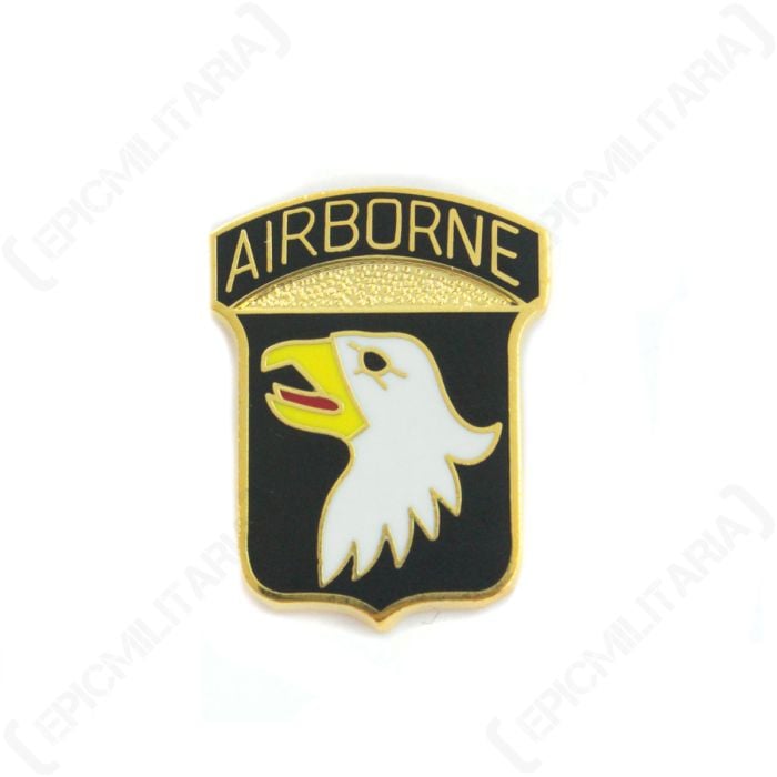 Front of shield-shaped 101st Airborne Pin Badge showing right facing, white eagle head with black eye, yellow beak, and red tongue detail, on black background. On top of the shield is a black arch with AIRBORNE in gold