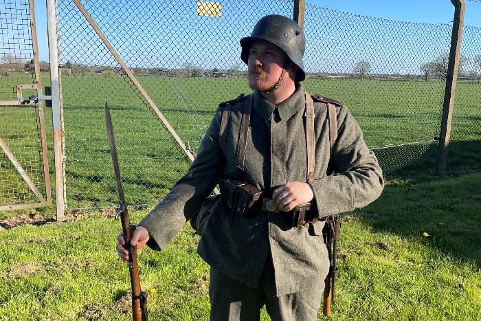 Dressed as a WW1 German soldier, a reenactor poses with helmet, uniform and rifle in front of a fence
