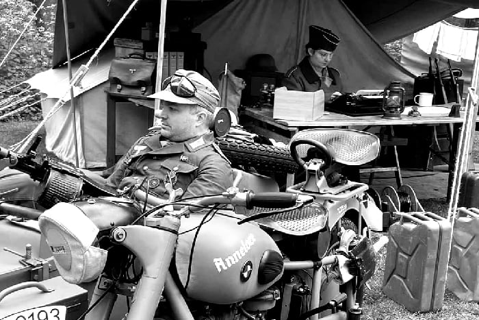 Black and white. In front a tent with desk and soldier using a typewriter, a motorcycle with sidecar hosts a soldier with a machine gun