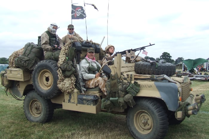 A land rover painted black and tan with SAS renactors inside, featuring lots of additional bags and camouflage netting.