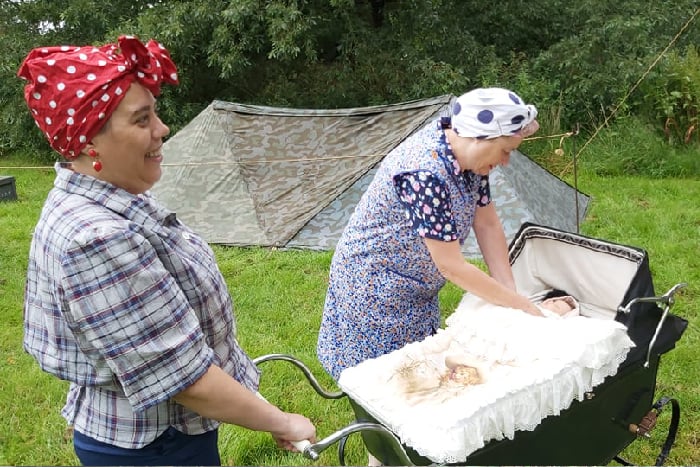 Two ladies in WW2 era clothing care for a baby in a pram in front of a campsite
