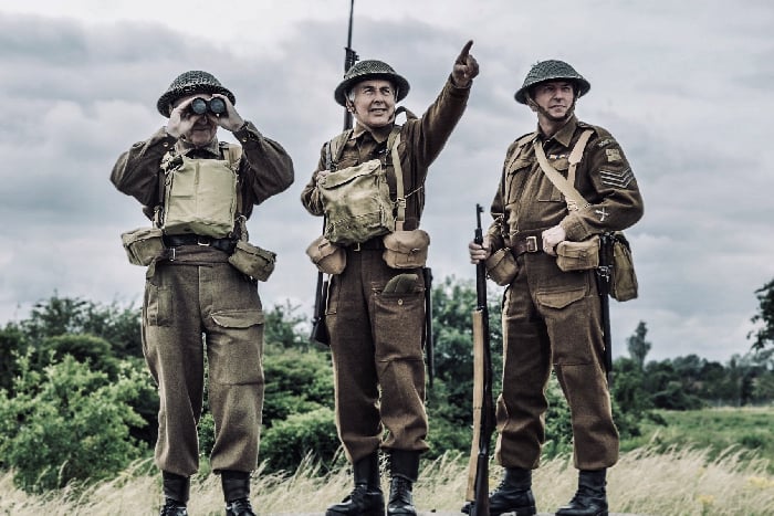 3 reenactors in WW2 British home gaurd uniforms with bags helmets and binoculars standing in front of trees and sky pointing into the distance