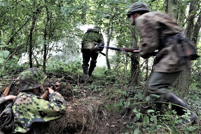 camouflaged ww2 german reenactors spring out from cover and head out of the bushes, away from the camera