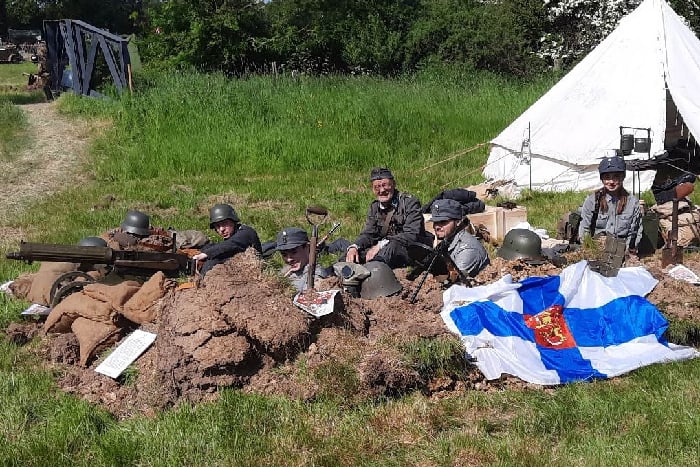 Members of the group behind a trench into the grass field, with machine guns and a finnish flag. Behind, a tent and hedgerow