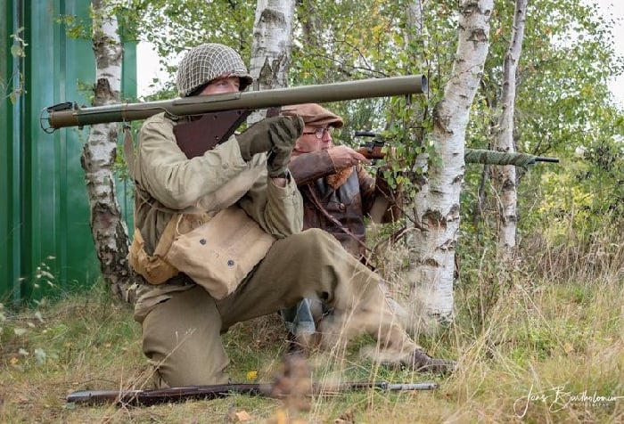 One reenactor aims a bazooka while another behind him aims a sniper rifle among trees
